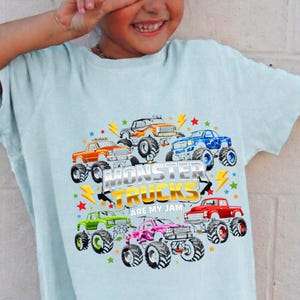 May include: Light blue t-shirt featuring a colorful graphic of monster trucks in various colors, including orange, blue, and pink. The text "MONSTER TRUCKS ARE MY JAM" is prominently displayed. The shirt is worn by a smiling child.