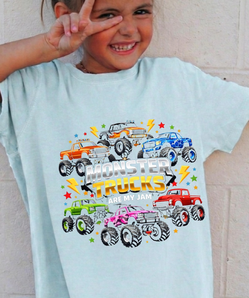 May include: Light blue t-shirt featuring a colorful graphic of monster trucks in various colors, including orange, blue, and pink. The text "MONSTER TRUCKS ARE MY JAM" is prominently displayed. The shirt is worn by a smiling child.