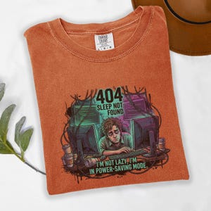 May include: Burnt orange Comfort Colors t-shirt with a graphic featuring a person at a computer with the text "404 Sleep Not Found" and "I'm Not Lazy, I'm in Power-Saving Mode."