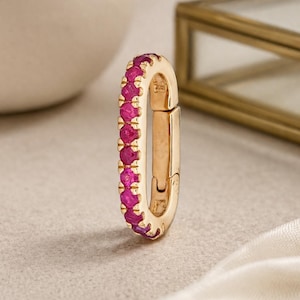 May include: A gold oval clasp, set with a line of bright pink gemstones. The clasp is open, showing the inner workings. The background features a white ceramic vase, a gold-framed box, and white fabric.