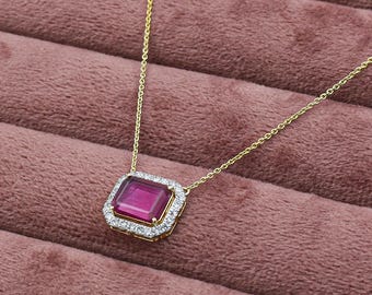 May include: A gold necklace with a rectangular, magenta-colored gemstone pendant. The pendant is surrounded by a halo of small, clear gemstones. The necklace is displayed on a pink velvet surface.
