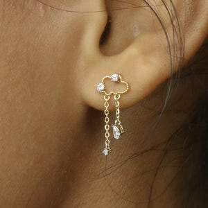 Può includere: Un orecchino a forma di nuvola color oro con tre piccole pietre trasparenti. L'orecchino ha una catena con una pietra trasparente e un ciondolo a forma di stella. L'orecchino è indossato sull'orecchio.