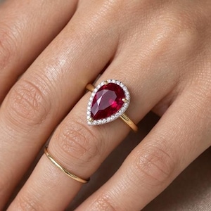 Puede incluir: Un anillo de oro con una gran piedra roja en forma de pera rodeada por un halo de pequeñas piedras blancas, acompañado de una banda dorada fina y sencilla en el dedo adyacente. La joya presenta un acabado en oro amarillo pulido.