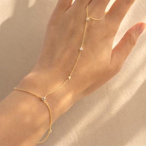 Puede incluir: Una delicada pulsera de cadena de mano dorada con pequeñas gemas brillantes. La pulsera se conecta a un anillo y cae elegantemente sobre el dorso de la mano. La joya se exhibe sobre un fondo de tonos claros.