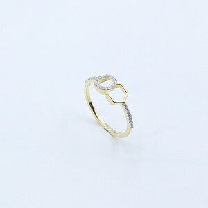 May include: A gold-colored ring with a geometric design. The ring features two connected shapes: a square and a hexagon. Small, clear stones are set along the band and in the square shape.