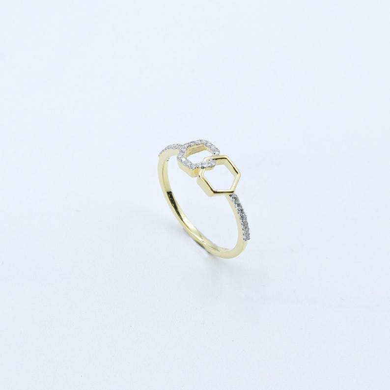 May include: A gold-colored ring with a geometric design. The ring features two connected shapes: a square and a hexagon. Small, clear stones are set along the band and in the square shape.