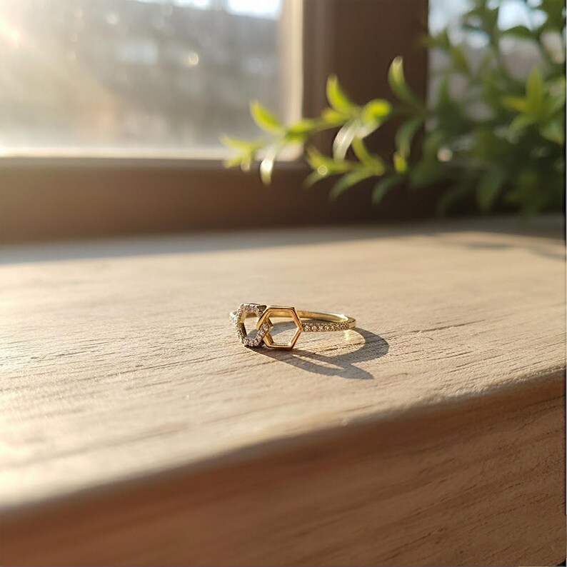 May include: A gold ring with a geometric design, featuring a heart and a hexagon shape, adorned with small, clear gemstones. The ring is set on a wooden surface, with sunlight illuminating the details. A piece of jewelry.