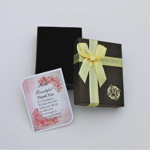 May include: A brown gift box tied with a yellow ribbon and bow, next to a black box and a card with pink floral accents. The card reads "Hello Beautiful Thank You". The gift box has a gold emblem.