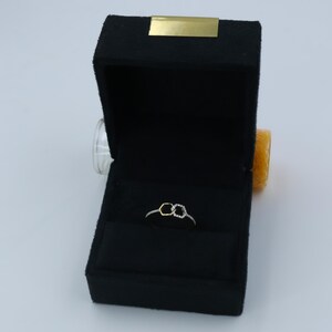 May include: A delicate ring with a double hexagon design, set in a black velvet jewelry box. The ring features a gold-colored hexagon and a silver-colored hexagon, both adorned with small, sparkling stones. The box has a gold-colored accent on the lid.