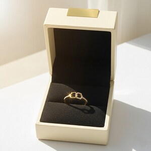 May include: A gold ring with a double-hexagon design, encrusted with small, clear gemstones, rests inside an open, cream-colored jewelry box with a black velvet interior. The box has a gold-colored rectangular accent on the lid.