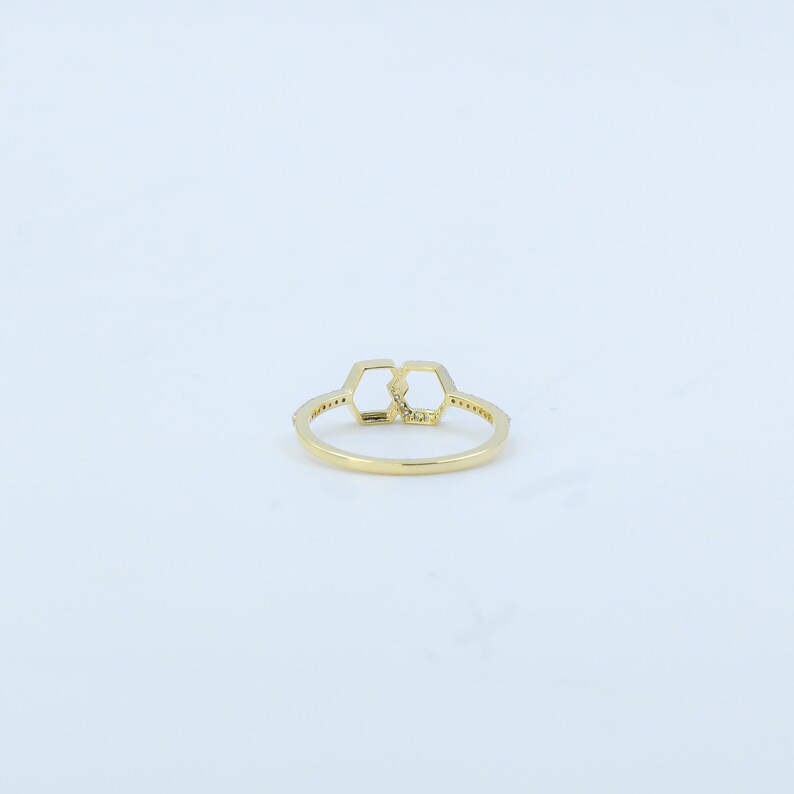 May include: A gold ring featuring a double hexagon design. The ring is adorned with small, sparkling stones along the band and within the geometric shapes. The ring is set against a white background.