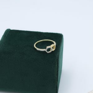 May include: A gold-colored ring with a double hexagon design. One hexagon is encrusted with small, clear gemstones, while the other is solid gold. The ring is set on a dark green velvet surface.