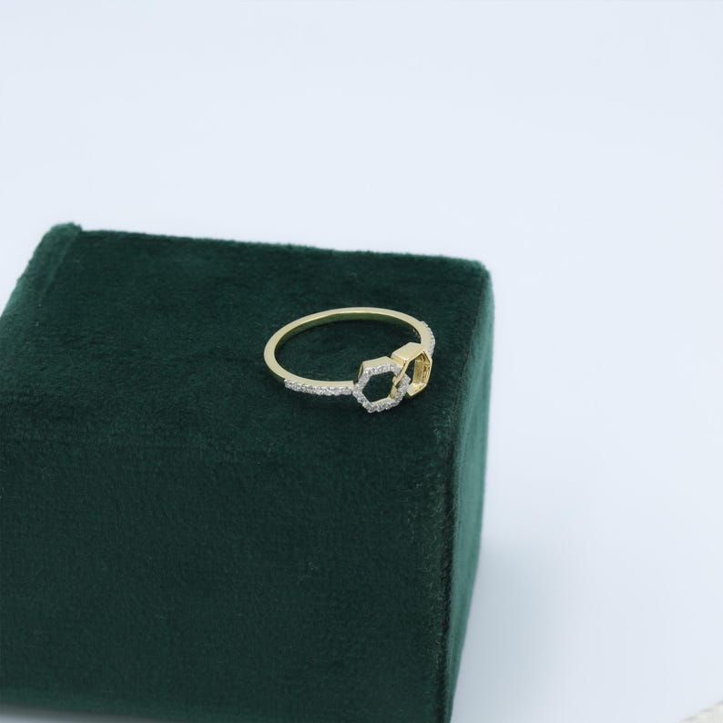 May include: A gold-colored ring with a double hexagon design. One hexagon is encrusted with small, clear gemstones, while the other is solid gold. The ring is set on a dark green velvet surface.