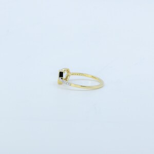 May include: A gold ring with a rectangular black stone as the centerpiece. The band is thin and features small, clear stones along one side. The ring is set against a white background, highlighting its design.