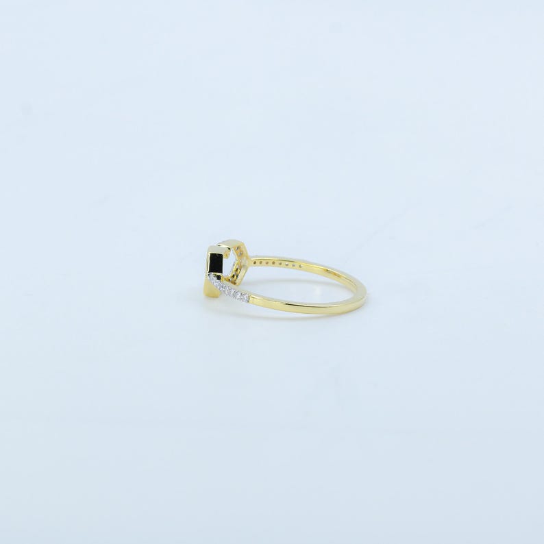 May include: A gold ring with a rectangular black stone as the centerpiece. The band is thin and features small, clear stones along one side. The ring is set against a white background, highlighting its design.
