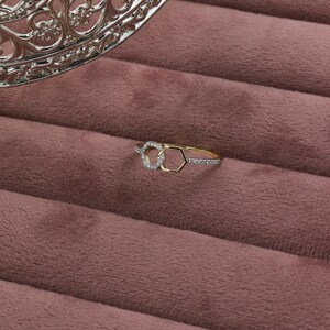 May include: A gold ring with a unique design featuring two interlocking shapes, one a circle and the other a hexagon, both adorned with small, sparkling stones. The ring rests on a pink velvet surface, with a decorative silver element in the background.