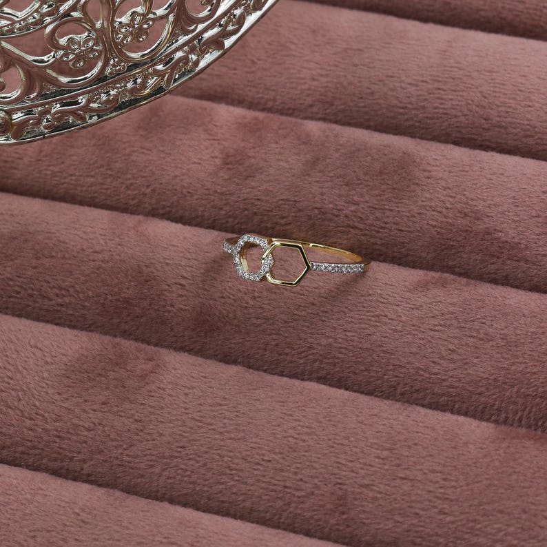 May include: A gold ring with a unique design featuring two interlocking shapes, one a circle and the other a hexagon, both adorned with small, sparkling stones. The ring rests on a pink velvet surface, with a decorative silver element in the background.
