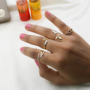May include: A hand displaying five gold-toned rings. The rings feature various designs, including a fish tail, a geometric shape, and bands with small, clear stones. The background includes colorful drinks in glass tubes.