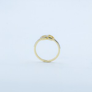 May include: A gold-colored ring with a knot design. The ring features small, clear gemstones along the band and on the knot. The ring is set against a white background, highlighting its details.