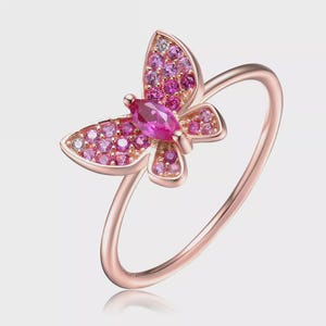May include: Rose gold butterfly ring with pink and purple gemstones. The butterfly design features a large marquise-cut pink gemstone in the center and smaller stones on the wings. The ring band is a delicate, thin design.