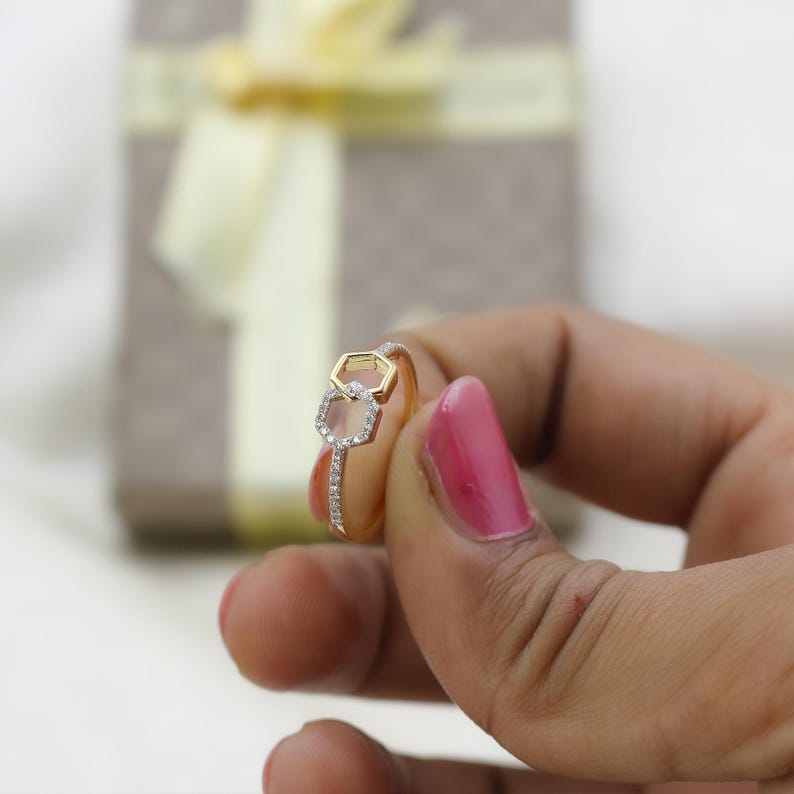 May include: A gold ring with a double hexagon design, one in gold and the other outlined with small, clear stones. The ring band is also adorned with small stones. The ring is held in a hand with pink nail polish.