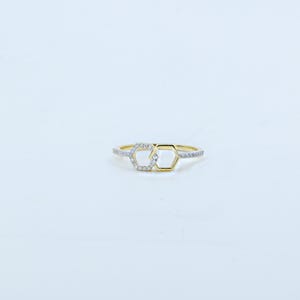 May include: A gold-colored ring featuring two interconnected hexagonal shapes. One hexagon is outlined with gold, while the other is filled with small, sparkling stones. The band is also adorned with similar stones, creating a delicate and elegant design.