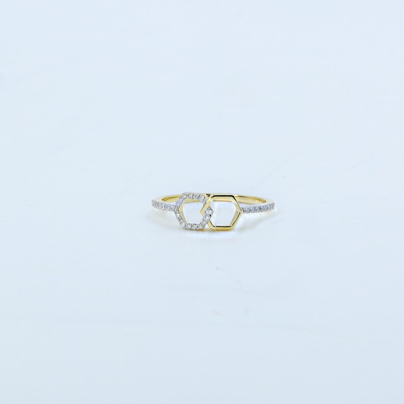 May include: A gold-colored ring featuring two interconnected hexagonal shapes. One hexagon is outlined with gold, while the other is filled with small, sparkling stones. The band is also adorned with similar stones, creating a delicate and elegant design.