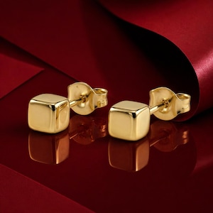 May include: A pair of gold cube stud earrings. The earrings are made of polished gold and have a simple, geometric design. The earrings are displayed on a red surface.