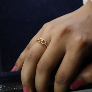 May include: A gold-toned ring with a double-loop design, one loop is encrusted with small, clear gemstones. The ring is on a finger, set against a dark background. The ring's design is delicate and elegant.