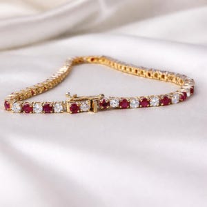 Puede incluir: Una pulsera de tenis dorada con piedras preciosas rojas y claras alternas. La pulsera se muestra sobre una superficie de satén blanco, con el cierre visible. Las piedras están espaciadas uniformemente.
