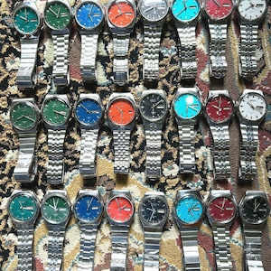 Puede incluir: Una colección de veinticuatro relojes de pulsera analógicos dispuestos en tres filas. Cada reloj cuenta con una esfera redonda con ventana de día y fecha, una correa de eslabones de metal plateado y una esfera vibrante en verde, azul, naranja, negro, rojo o blanco.