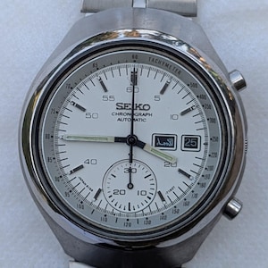 Op de afbeelding: Een zilveren Seiko Chronograph Automatic horloge met een witte wijzerplaat. Het horloge heeft een tachymeter, meerdere sub-wijzerplaten en de datum. De horlogeband is zilverkleurig en van metaal. De wijzerplaat toont de tekst "SEIKO CHRONOGRAPH AUTOMATIC".
