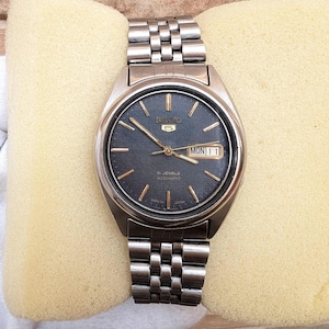 May include: A silver-tone stainless steel Seiko 5 automatic wristwatch with a textured dark blue dial, gold-tone hands and hour markers, and a day-date window at the three o'clock position. The watch features a metal link bracelet.