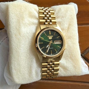 Op de afbeelding: Een goudkleurig Seiko 5 automatisch polshorloge met een ronde, diepgroene wijzerplaat met zonnestraalpatroon, gouden uurmarkeringen en wijzers. Het horloge heeft een dag-datumvenster op de drie-uurpositie en een bijpassende metalen schakelarmband.