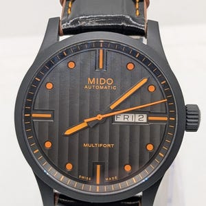 Puede incluir: Reloj automático Mido Multifort negro con detalles en naranja. El reloj presenta una esfera negra con rayas verticales, agujas e indicadores horarios naranjas. Correa de cuero negro con costuras naranjas y caja de acero inoxidable.