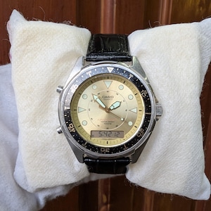 May include: A Casio Quartz analog-digital watch with a gold-toned circular dial, luminous hands, and a black rotating bezel. The watch features a digital display at the bottom, a stainless steel case, and a black textured leather strap.