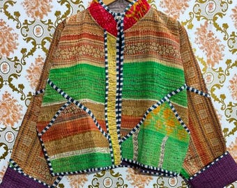 May include: A colorful patchwork jacket with a mandarin collar. The jacket features horizontal stripes of green, orange, and brown fabrics, accented with black and white trim. The cuffs are a deep purple. The jacket is hanging on a wooden hanger.