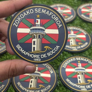 Pode incluir: Remendo circular azul marinho com um design de farol, o texto "ZOKOAKO SEMAFOROA" e "SEMAPHORE DE SOCOA 1863". O remendo tem um design de bandeira vermelha, branca e verde no centro.