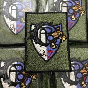 Puede incluir: Una colección de parches bordados en verde oliva. Cada parche presenta un diseño de escudo con las letras estilizadas "A" y "B", un tigre rugiente y el número "7". Los parches están enmarcados con un borde negro.