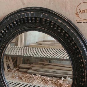 Pu&ograve; includere: Una cornice per specchio ovale nera con dettagli decorativi. La cornice ha una finitura anticata, che rivela tocchi di legno. L'immagine mostra anche altre cornici in legno non finite e un'ambientazione da officina.