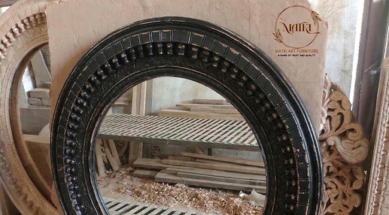 Pu&ograve; includere: Una cornice per specchio ovale nera con dettagli decorativi. La cornice ha una finitura anticata, che rivela tocchi di legno. L'immagine mostra anche altre cornici in legno non finite e un'ambientazione da officina.