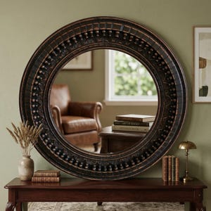 Może przedstawiać: Duże, okrągłe, ciemnobrązowe lustro z ozdobną, rzeźbioną ramą. Lustro odbija pokój ze skórzanym fotelem, książkami i małym stolikiem. Lustro jest umieszczone na ciemnym drewnianym stole.