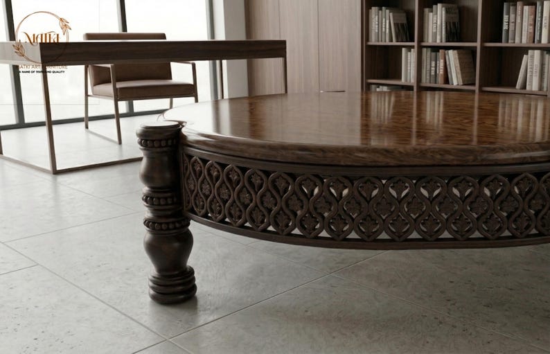 May include: A large, round, dark brown wooden coffee table with a glossy finish. The table features intricate carved detailing around the base and a decorative, turned leg. The table is in a room with a desk, chair, and bookshelves.