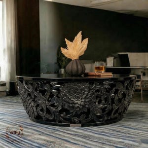 Puede incluir: Una taza de cerámica blanca con un asa negra y el texto "But First, Coffee" en negrita negra. La taza se encuentra sobre un fondo borroso, sugiriendo un ambiente acogedor.