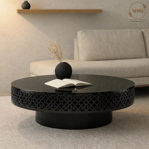 May include: A black round coffee table with a decorative patterned edge. A black vase with dried flowers and an open book sit on the table. A beige sofa is in the background. The table is a modern piece of furniture.