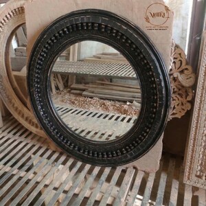 Pu&ograve; includere: Uno specchio rotondo nero con una cornice decorativa. La cornice presenta un motivo dettagliato di punti in rilievo e forme rettangolari. Lo specchio &egrave; posto su uno sfondo neutro, con altre cornici in legno visibili.