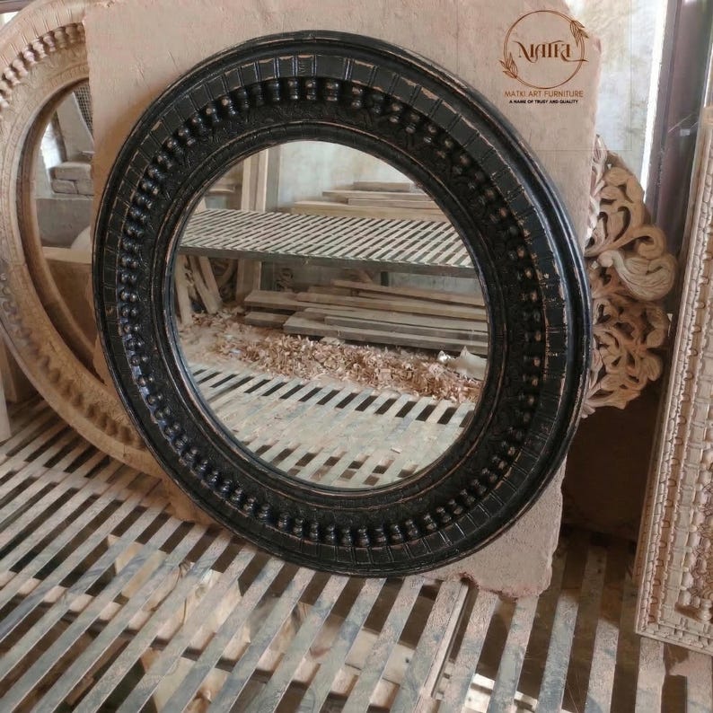 Pu&ograve; includere: Uno specchio rotondo nero con una cornice decorativa. La cornice presenta un motivo dettagliato di punti in rilievo e forme rettangolari. Lo specchio &egrave; posto su uno sfondo neutro, con altre cornici in legno visibili.