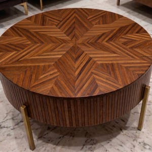 Puede incluir: Mesa de centro redonda de madera oscura con un diseño de estrella en la parte superior. La mesa tiene patas doradas y laterales con ranuras verticales. El diámetro de la mesa es de aproximadamente 100 cm.