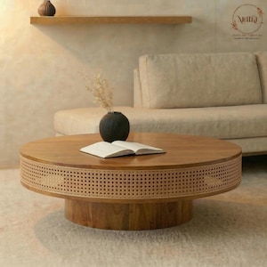 Puede incluir: Una mesa de centro redonda de madera con un borde de ratán tejido. La mesa tiene una base de madera oscura y una tapa de madera clara. Un jarrón negro con flores secas y un libro abierto están sobre la mesa. La mesa está en un salón.