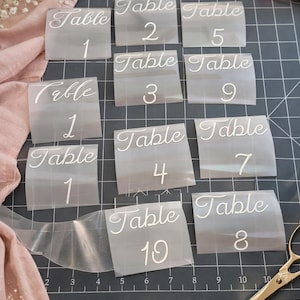 Peut inclure: Autocollants de numéros de table en vinyle transparent avec une police blanche. Chaque autocollant affiche le mot "Table" et un numéro de 1 à 10. Parfait pour les mariages ou événements. Les autocollants sont disposés sur un tapis de coupe gris foncé.